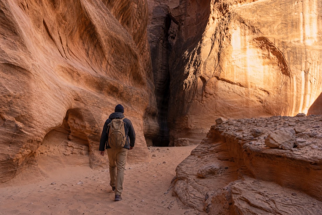 Warmly dressed traveler in layered jacket and beanie walking on sand toward a canyon entrance