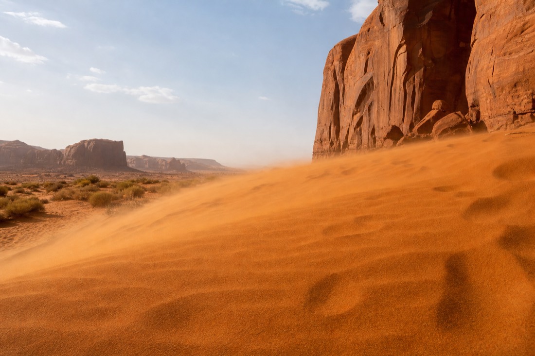 Wind lifting fine orange sand off desert dunes near sandstone cliffs on a clear spring day