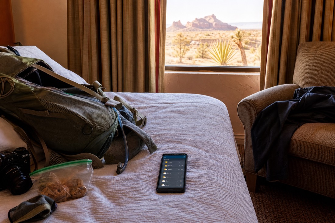 Traveler reviewing a weather forecast on a phone beside a packed daypack in a hotel room
