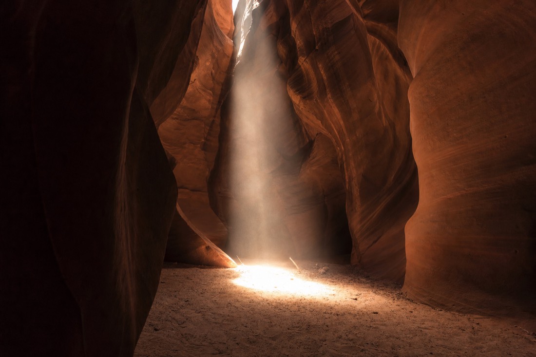 Midday light shaft descending through a slot canyon ceiling opening to the sandy floor