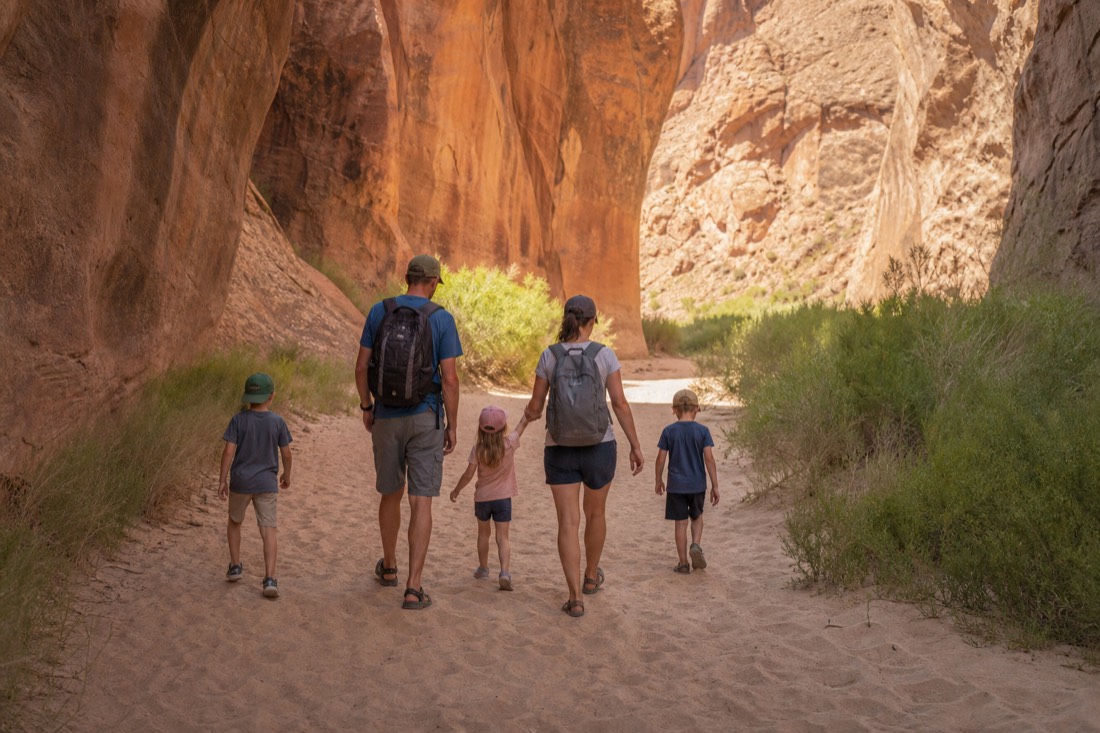 Family with children walking through soft deep sand toward a canyon entrance in bright sun