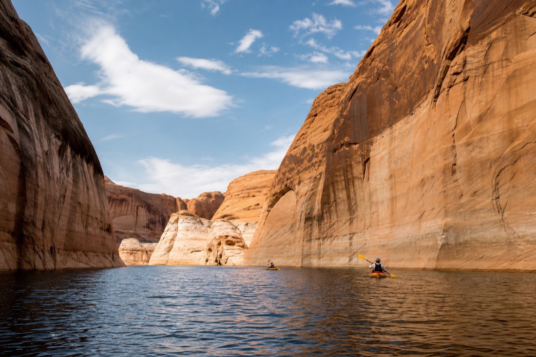 Kayakers paddling across calm blue water between tall sandstone canyon walls on Lake Powell