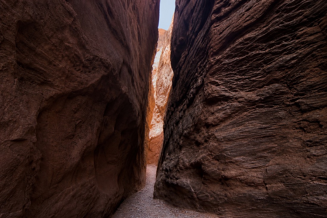 Narrow shadowed slot canyon passage with rough-textured walls and a sliver of sky above