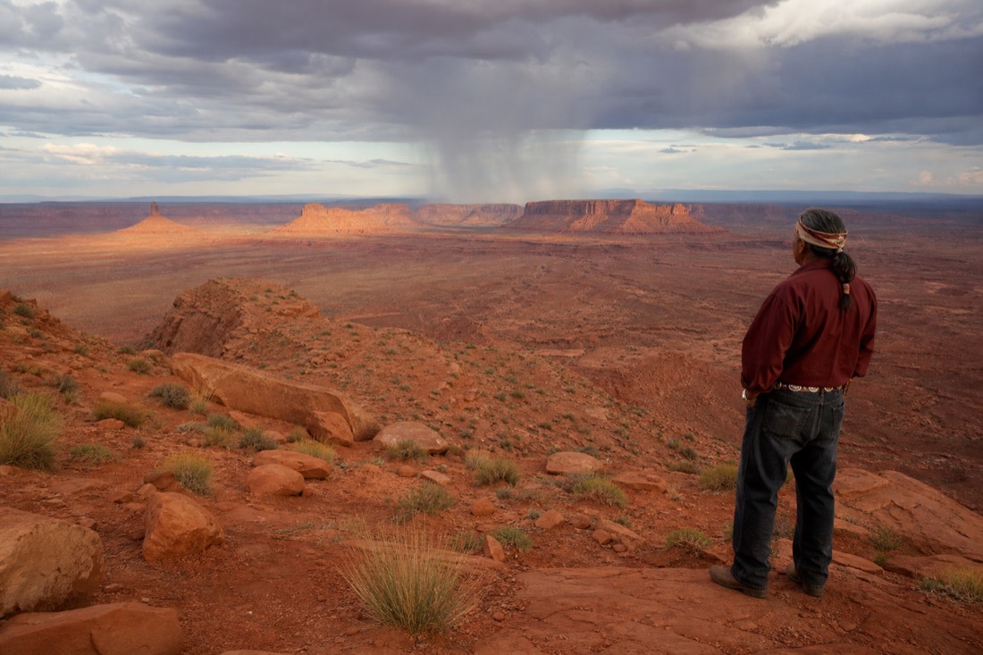 Navajo guide watching a distant afternoon rain curtain over red mesas from a dry desert overlook