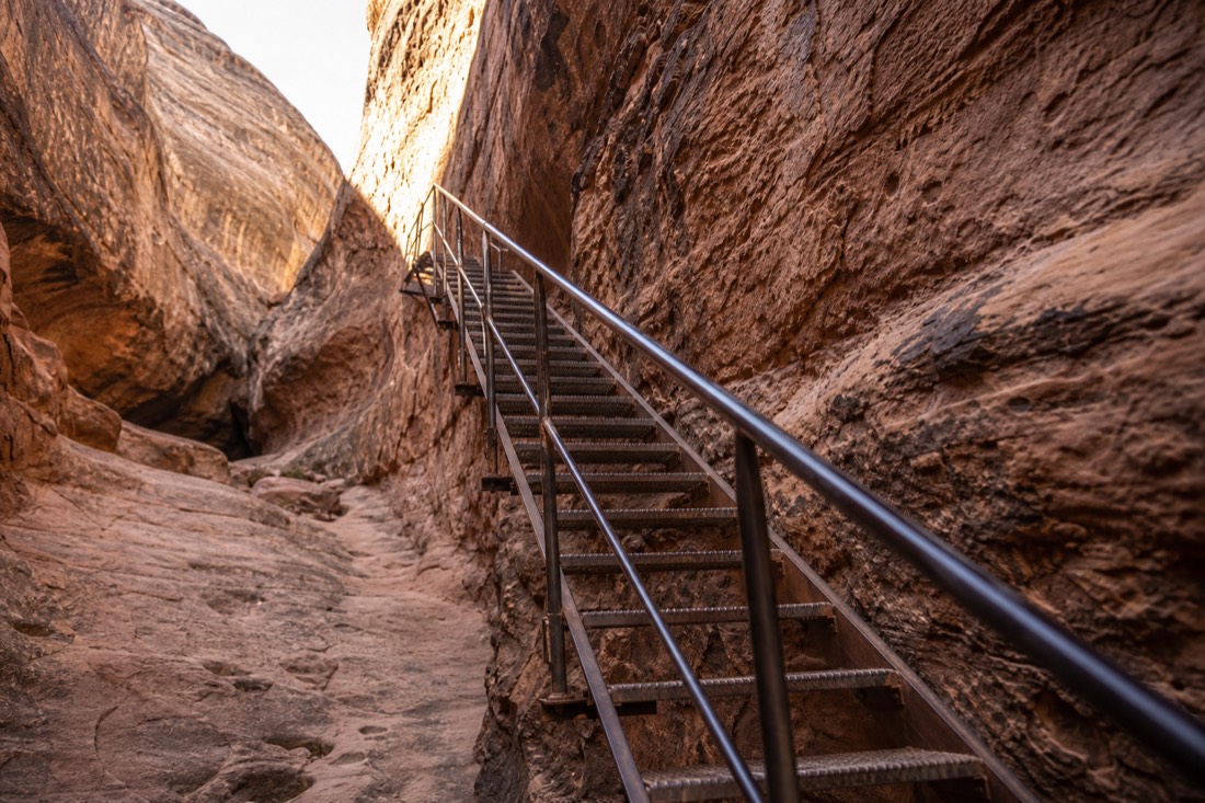 Metal stairway bolted to the sandstone wall of a slot canyon with a handrail catching morning light