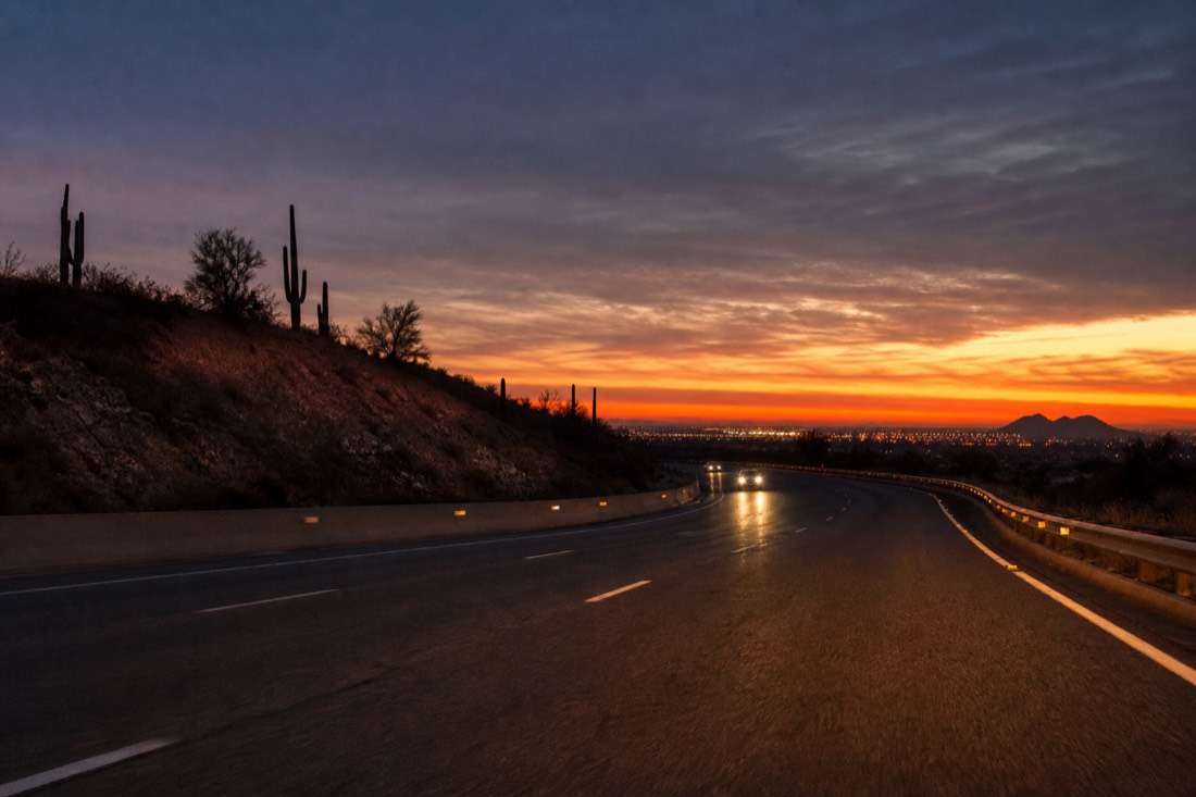 Car headlights on a dark interstate leaving a city before sunrise with saguaros silhouetted