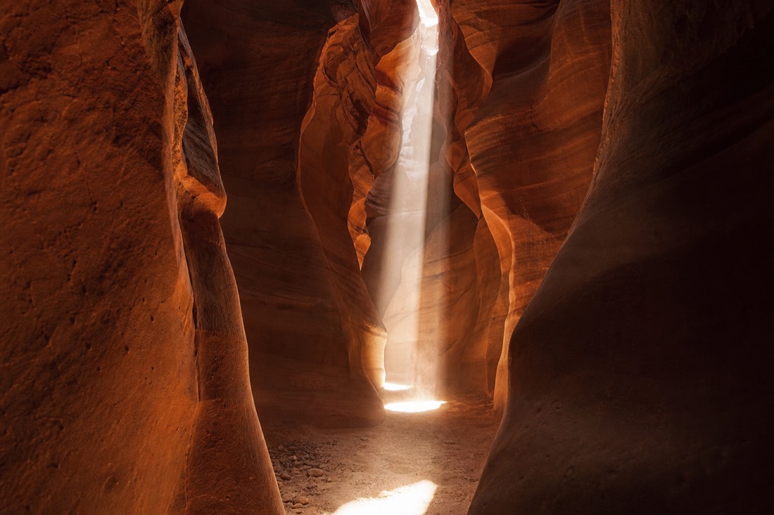 Faint morning glow on slot canyon walls giving way to a strong midday light column on the sand