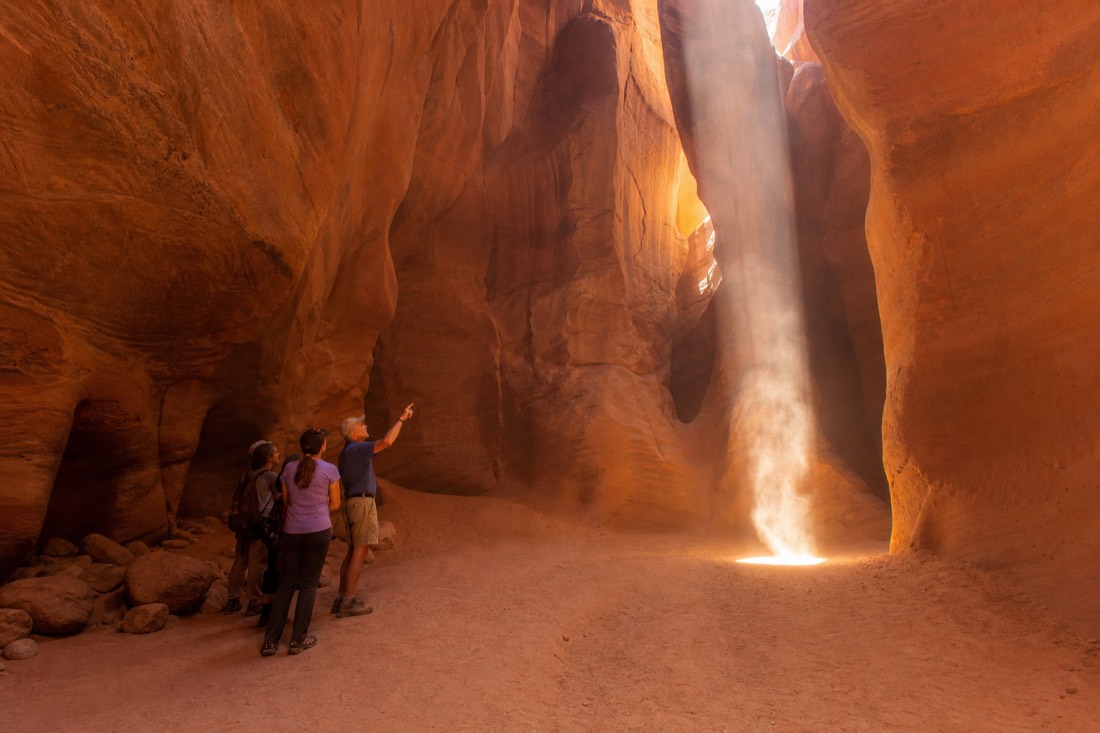Small group standing to one side of a slot canyon chamber as a guide gestures toward a light column