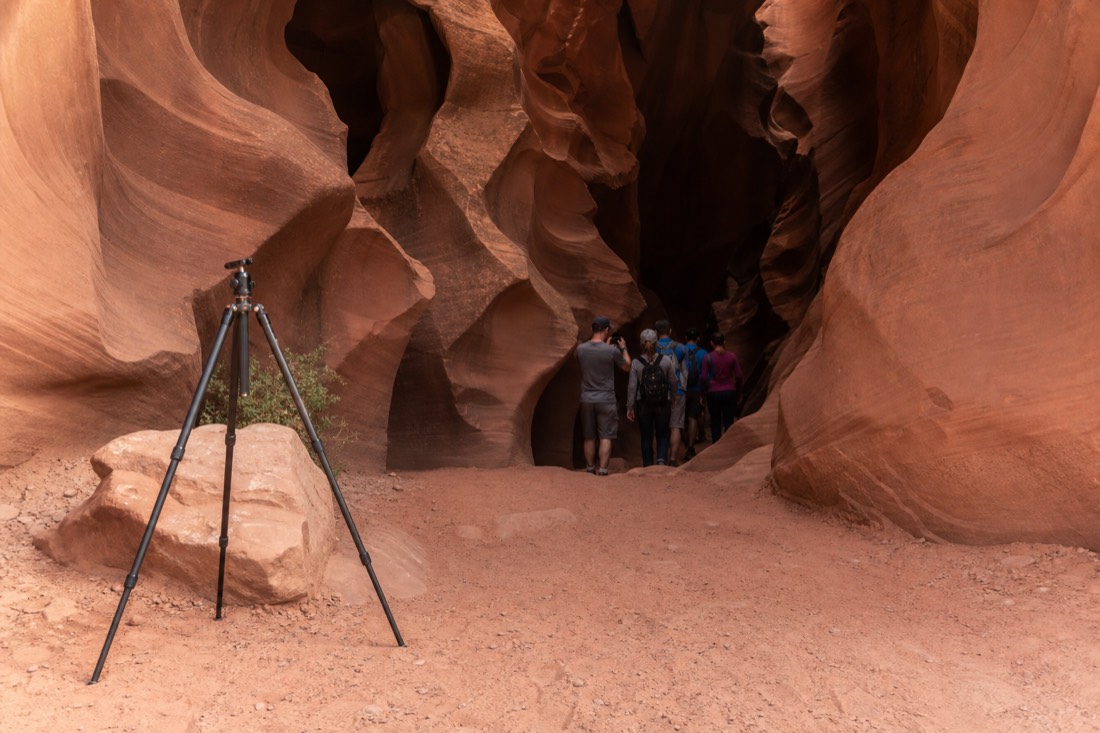 Old tripod left outside a slot canyon entrance while a guided group walks in with phones and handheld cameras