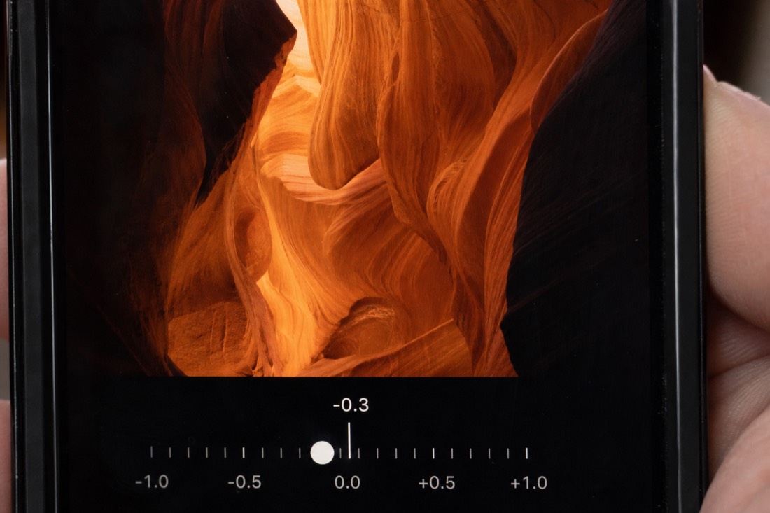 Close-up of a phone screen with exposure adjustment slider framing bright orange sandstone curves in a dim slot canyon