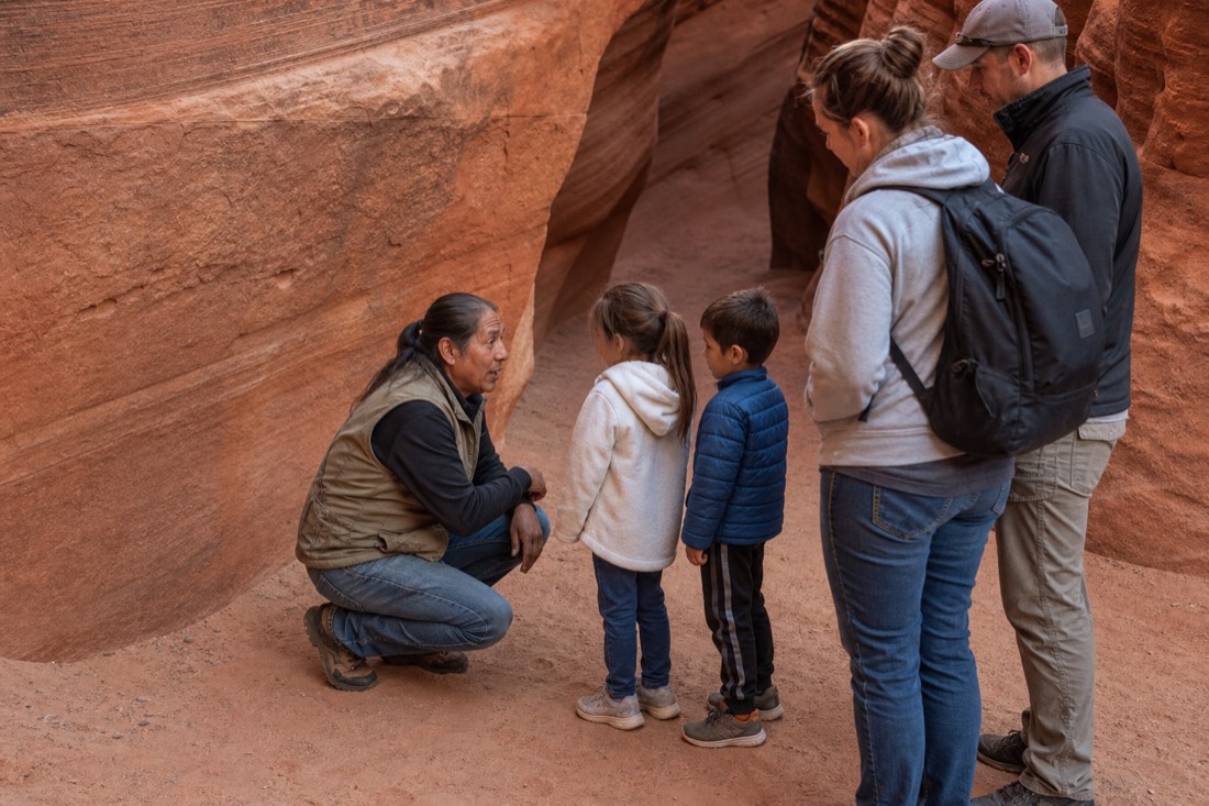 Navajo guide crouching to speak with two children at a slot canyon entrance while parents watch in the early morning