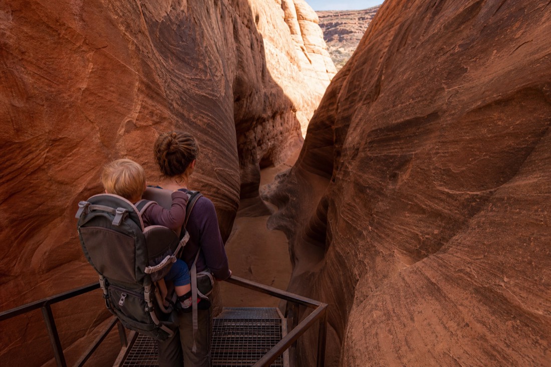 Parent carrying a toddler in a hiking backpack at the top of a steel stairway leading into a slot canyon