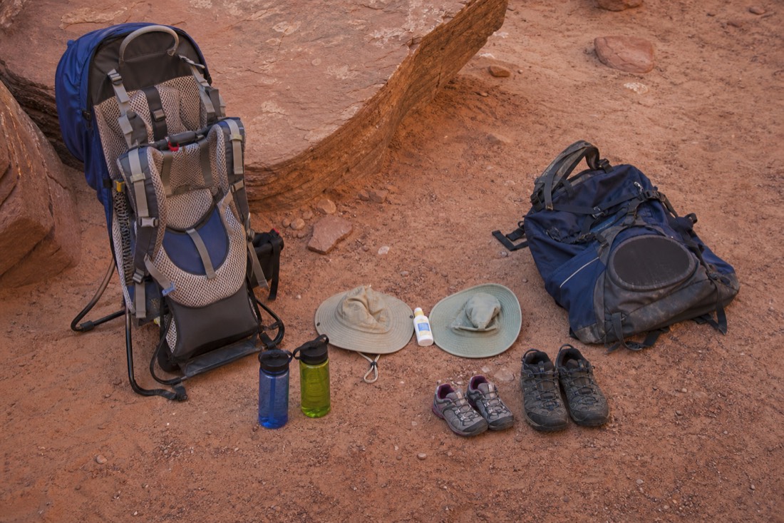 Flat lay of family canyon kit: child carrier, small water bottles, brimmed hats, sunscreen, and closed shoes, with a backpack set aside