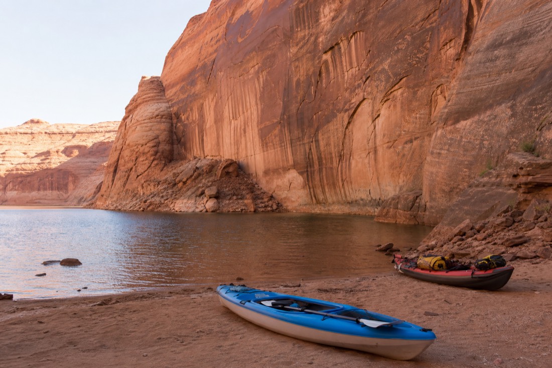 Kayaks pulled up on a sandy Lake Powell cove beneath tall sandstone walls in afternoon light