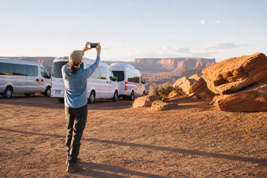 Traveler holding up a phone in a bright desert parking area with tour vehicles waiting behind