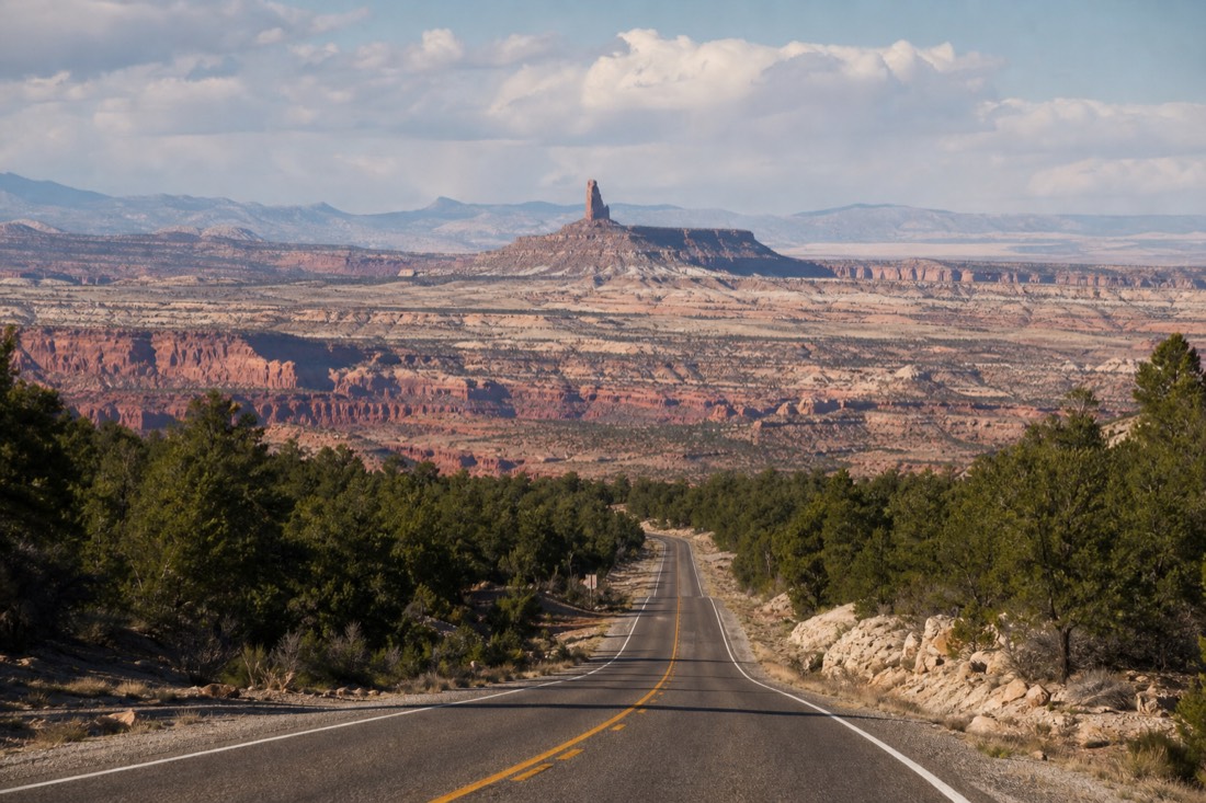 Two-lane highway descending from pine forest toward painted desert flats with a stone watchtower silhouette behind