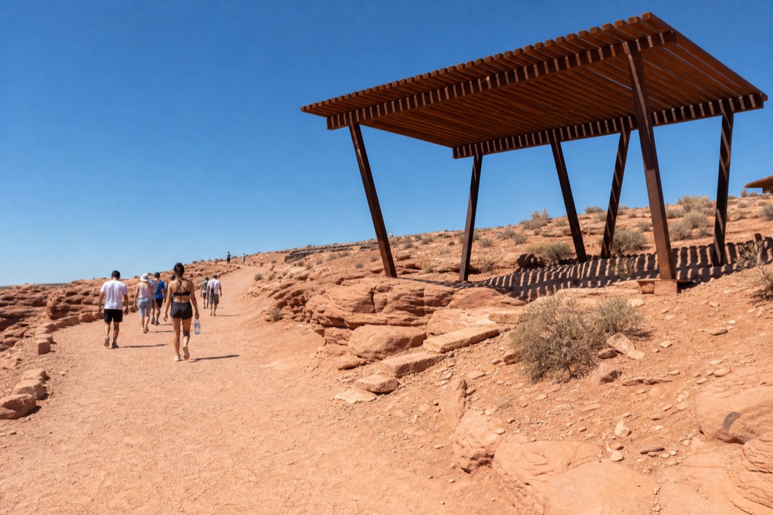 Shade structure beside the packed dirt Horseshoe Bend trail with hikers carrying water bottles under a hard blue sky