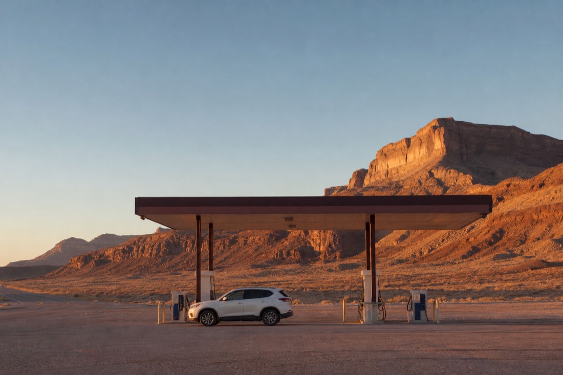 Lone car at a desert fuel station at dawn with empty highway and sandstone bluffs behind