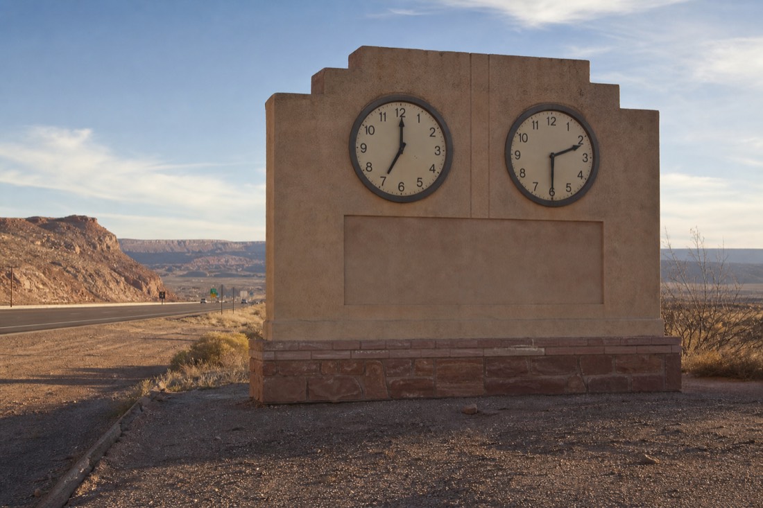 Roadside sign at a state line on a desert highway with two clocks showing different times