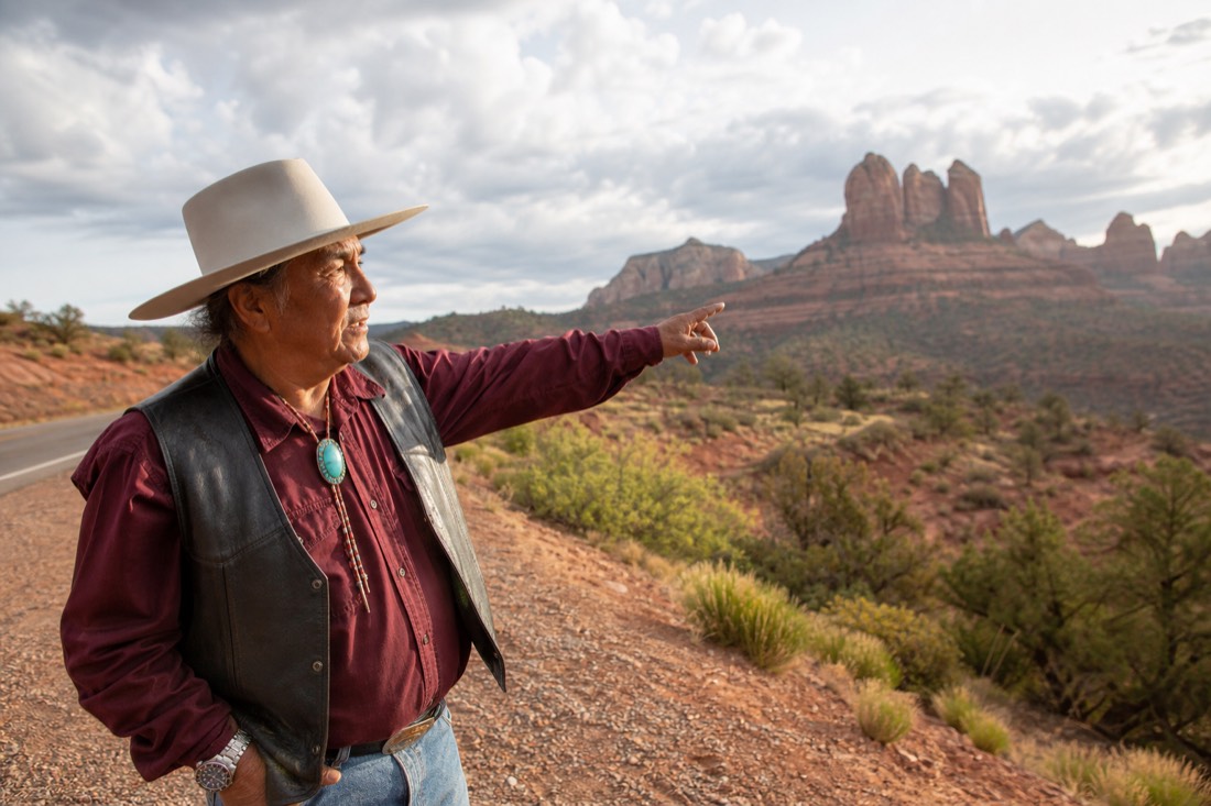 Navajo guide in a wide-brim hat gesturing toward red sandstone country from a roadside pullout