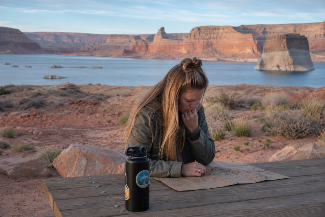 Traveler studying a hand-drawn itinerary at a picnic table with Lake Powell and red cliffs behind