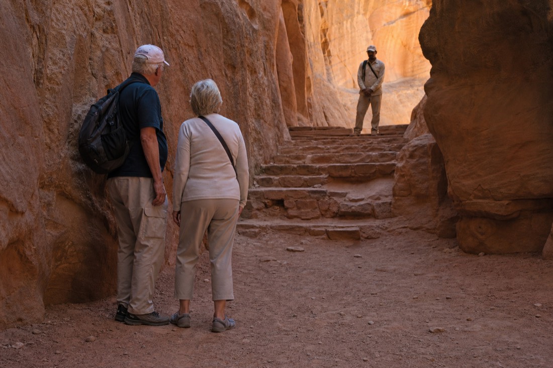 Older couple pausing at the shaded base of a slot canyon staircase while a guide waits ahead