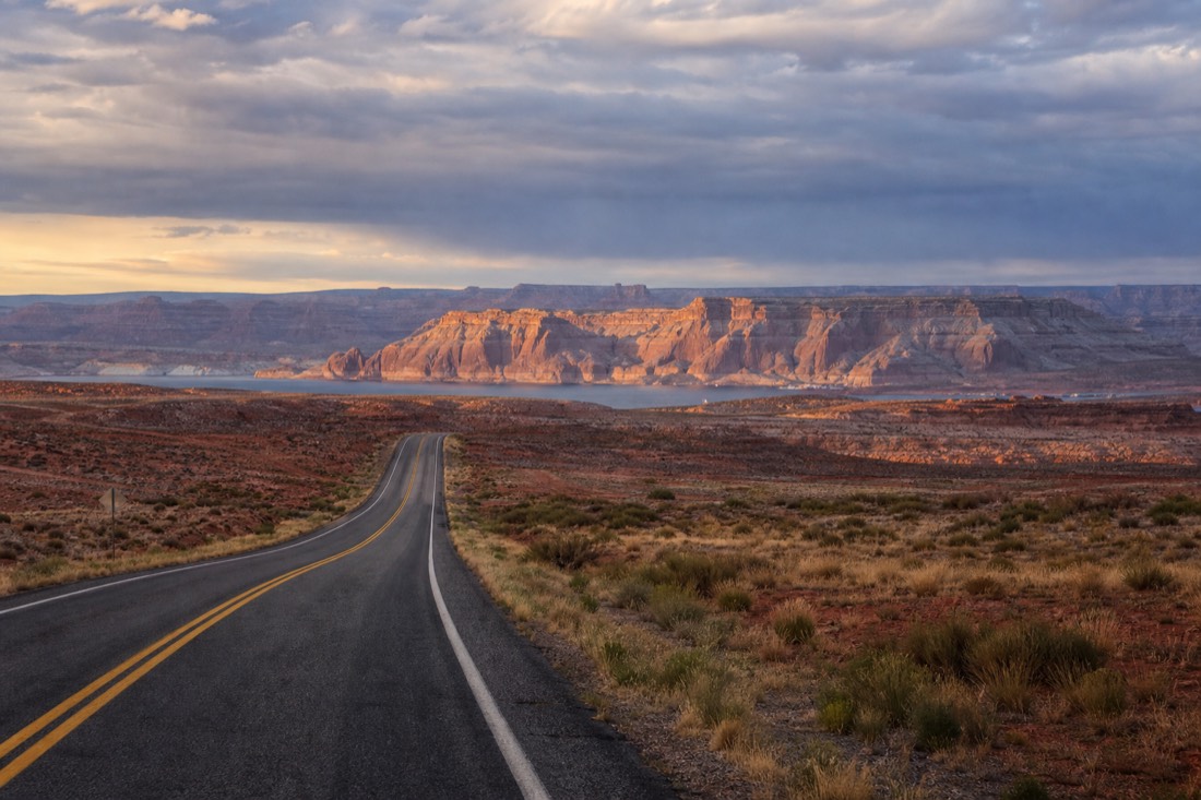 Two-lane desert highway approaching Page, Arizona at sunrise with red mesas on the horizon