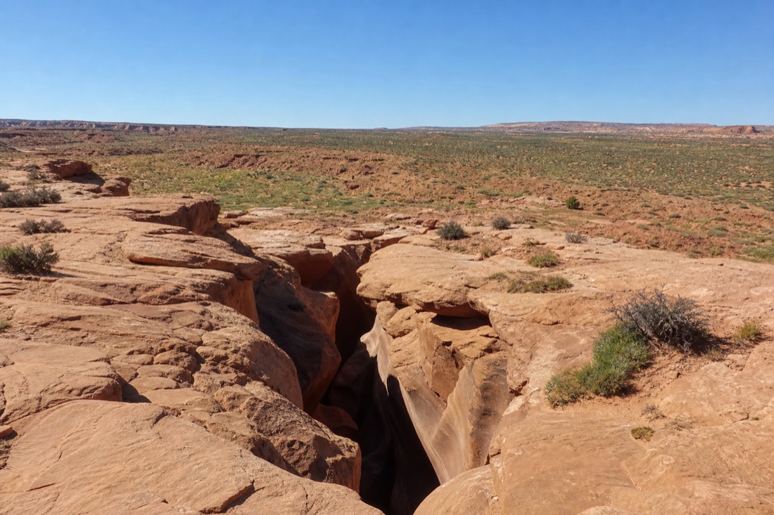 Slot canyon rim under a clear spring sky with a light dusting of green on the desert floor
