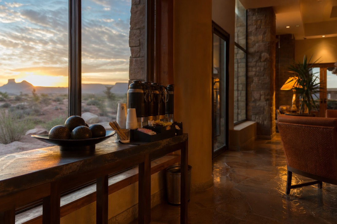 Hotel lobby coffee station at dawn with a traveler filling a to-go cup beside a window showing desert twilight