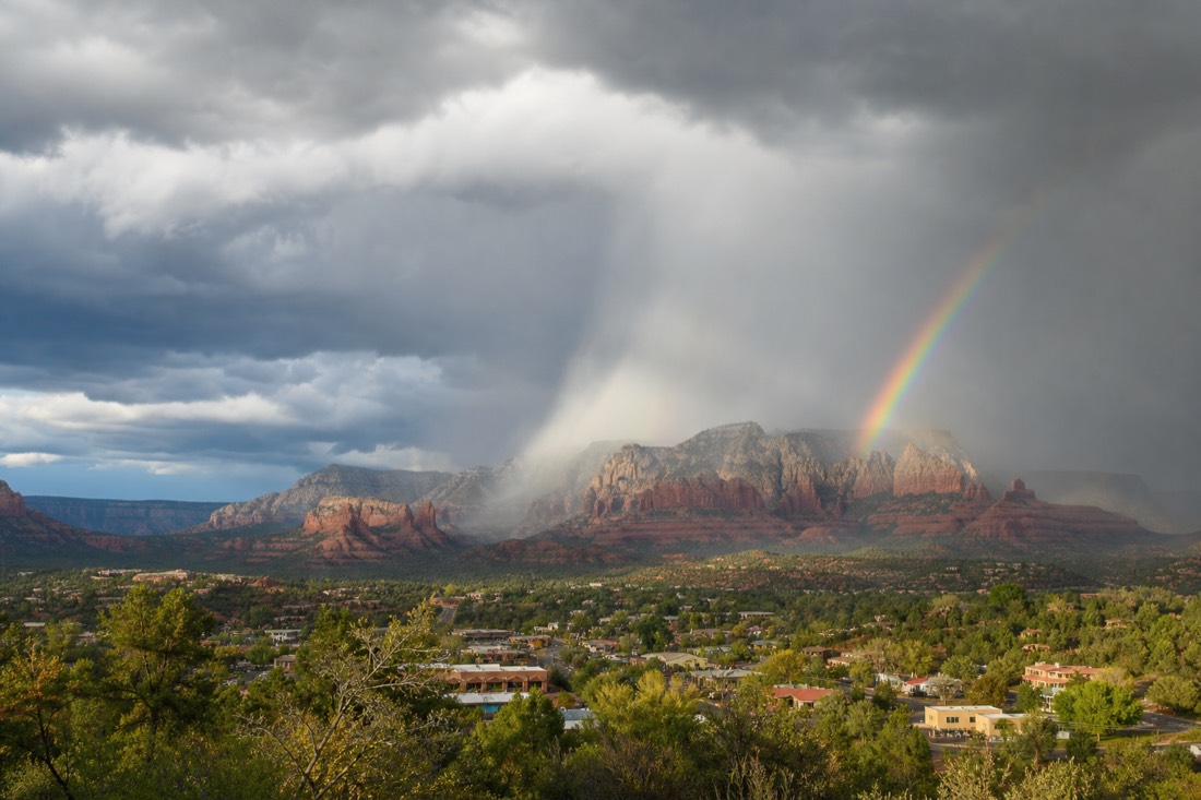 Dark monsoon storm cell dropping rain over red mesas behind a small town with a rainbow at its edge