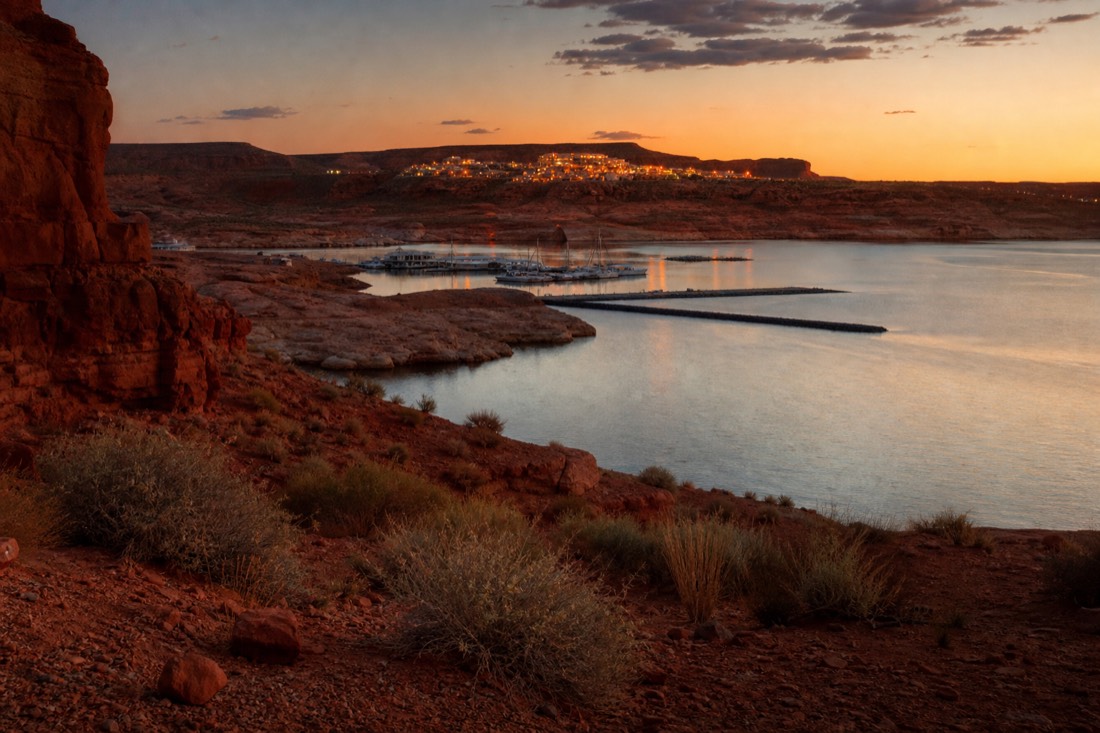 Sunset over a marina bay with red cliffs across the water and a small town glowing on the mesa in the distance
